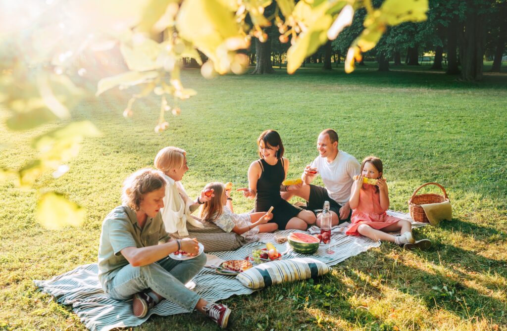 Big family sitting on the picnic blanket in the park on a sunny day. They are smiling, laughing and eating boiled corn and watermelon. 